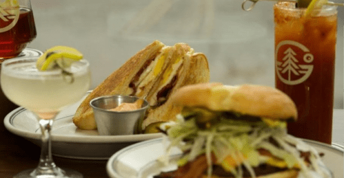 A close-up of a sandwich and a burger, accompanied by cocktails and garnishes on a wooden table.