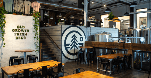 Interior of a brewery featuring wooden tables, a mural of a tree, and brewing equipment in a modern, industrial setting.