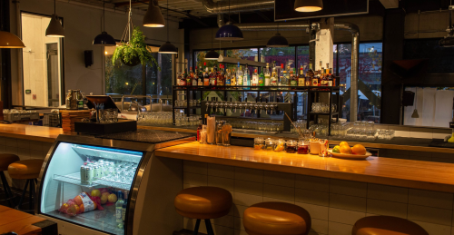 A cozy bar interior with a wooden counter, shelves of liquor, and a display of drinks and fruit. Warm lighting creates a welcoming atmosphere.