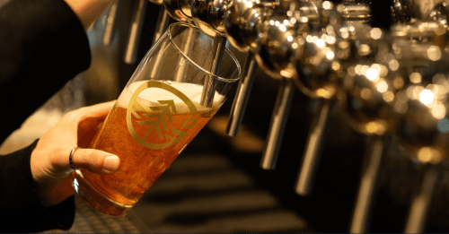 A hand pours a golden beer into a glass with a logo, from a row of shiny beer taps.