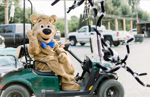 A bear mascot waves from a decorated golf cart, surrounded by Halloween-themed decorations.