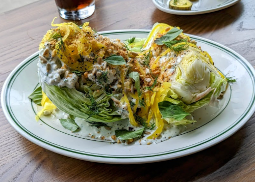 A plate of wedge salad topped with herbs, crunchy toppings, and a creamy dressing, served with a drink in the background.