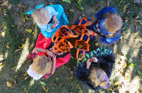 Four children in costumes sit on the ground, surrounded by orange bags filled with treats, enjoying their Halloween haul.