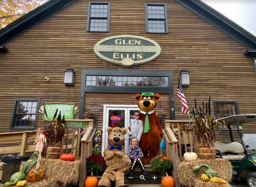 A family poses with two bear mascots outside a rustic building decorated for fall, featuring pumpkins and autumn decor.