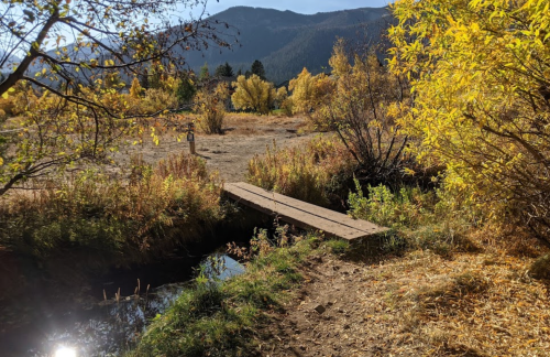 A wooden bridge crosses a small stream, surrounded by autumn foliage and mountains in the background.