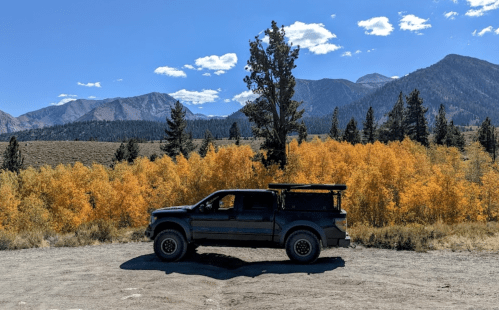 A black truck parked on a dirt road with vibrant autumn trees and mountains in the background under a clear blue sky.