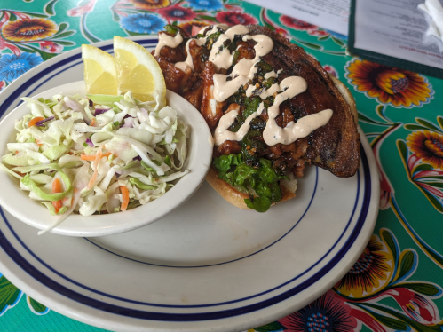 A grilled fish sandwich topped with sauce and greens, served with a side of coleslaw and lemon wedges on a colorful table.