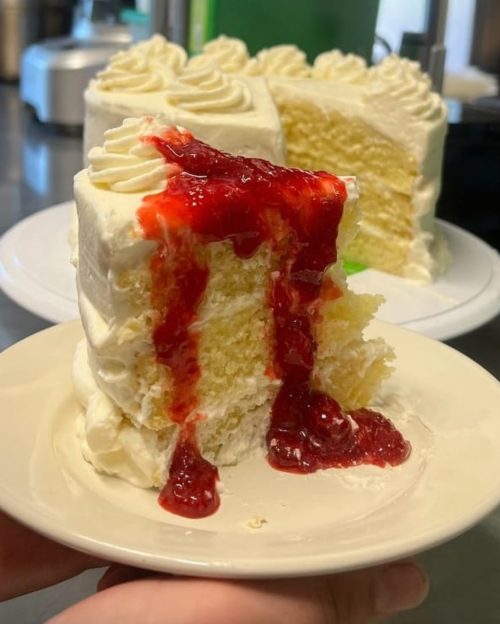 A slice of yellow cake with white frosting and strawberry sauce oozing from the center, served on a plate.