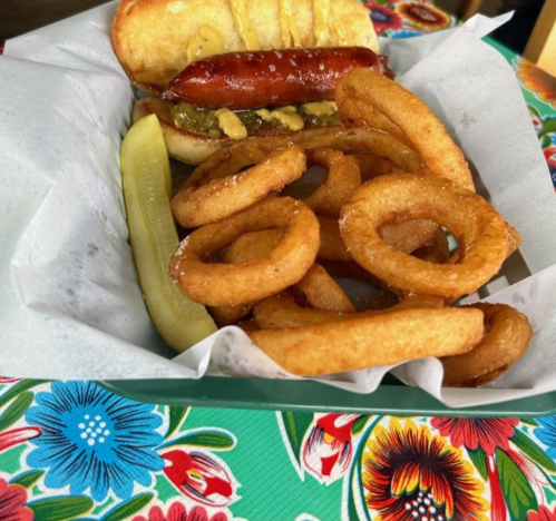 A hot dog in a bun with mustard and relish, served with crispy onion rings and a pickle slice on a colorful table.