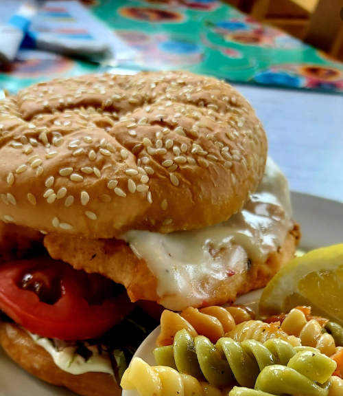 A sesame seed bun sandwich with fried chicken, lettuce, tomato, and sauce, served with a side of colorful pasta salad.