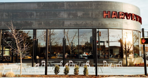 Modern restaurant exterior with large windows, "HARVEY'S" sign, outdoor seating, and decorative lights.