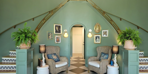 A stylish entryway with green walls, two wicker chairs, plants, and framed photos, leading to a restroom.