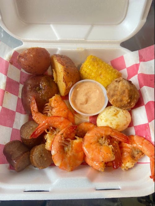 A takeout box filled with shrimp, potatoes, corn, sausage, and a dipping sauce.