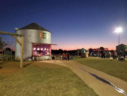 A rustic grain silo converted into a shop, with people lined up outside at dusk, under a colorful sunset sky.