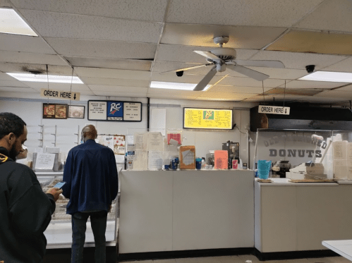 A donut shop interior with a counter, menu signs, and customers waiting to order. Ceiling fan visible.