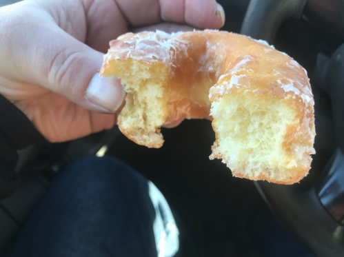 A hand holding a partially eaten glazed donut, with a car interior visible in the background.