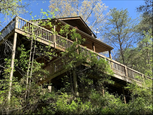 A wooden cabin nestled among trees, featuring multiple balconies and surrounded by lush greenery under a clear blue sky.