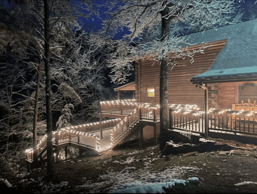 A cozy log cabin surrounded by snow, illuminated by warm lights on the deck and stairs at night.
