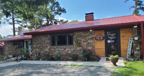 A rustic stone building with a red metal roof, surrounded by greenery and flowers, featuring a "Welcome" sign.