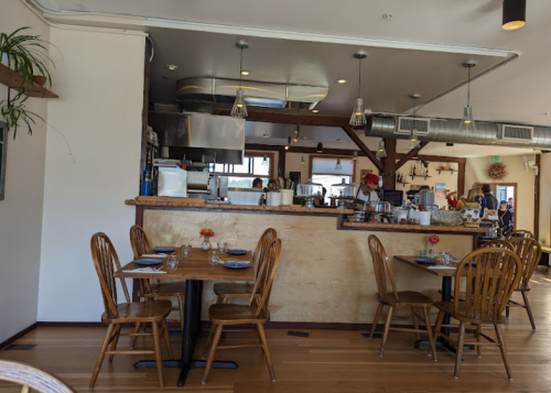 Cozy restaurant interior with wooden tables, a counter, and a kitchen area in the background. Bright and inviting atmosphere.