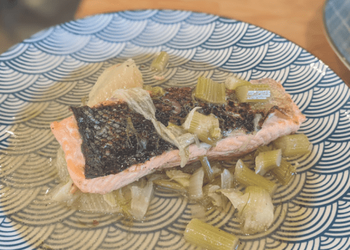 A piece of salmon with crispy skin, served on a patterned plate with sautéed celery and onions.