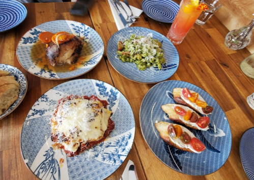A table set with four plates of colorful dishes, including pasta, meat, and appetizers, alongside drinks and utensils.
