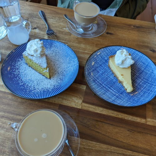 Two slices of cake topped with whipped cream on blue plates, accompanied by cups of coffee on a wooden table.