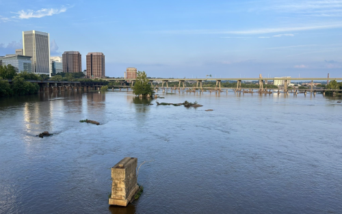 A calm river scene with city buildings in the background and a bridge crossing over, under a clear blue sky.