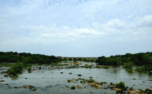 A serene river scene with a bridge in the background, surrounded by lush greenery and rocky outcrops under a cloudy sky.