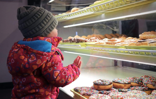 A child in a colorful coat and hat gazes at a display of donuts with sprinkles in a bakery.