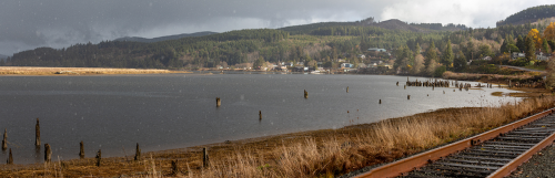 A tranquil landscape featuring a river, overcast skies, and a railway track alongside grassy banks and distant hills.