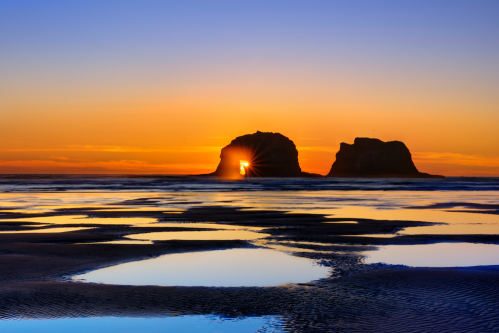 Sunset over two rock formations with reflections in the water, creating a serene coastal landscape.