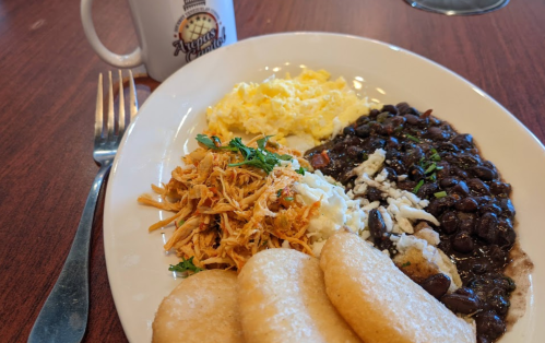 A plate of scrambled eggs, black beans, shredded chicken, cheese, and three arepas, with a coffee mug in the background.