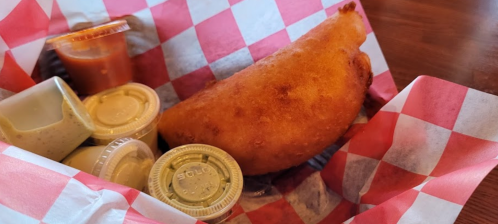 A golden-brown fried food item in a basket with red and white checkered paper, accompanied by various dipping sauces.