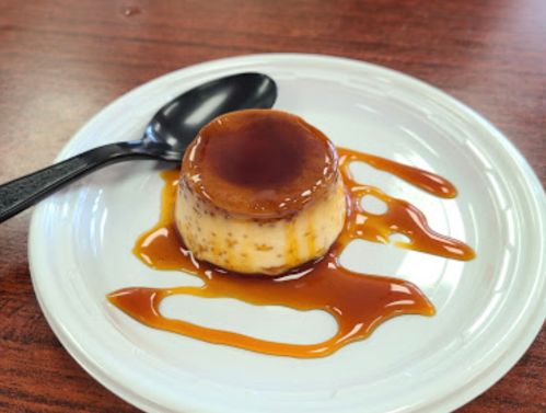 A creamy flan topped with caramel sauce on a white plate, accompanied by a black spoon.