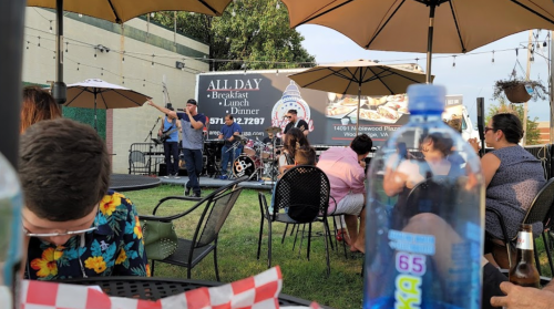 A lively outdoor scene with a band performing on stage, diners seated at tables under umbrellas, and a water bottle in focus.