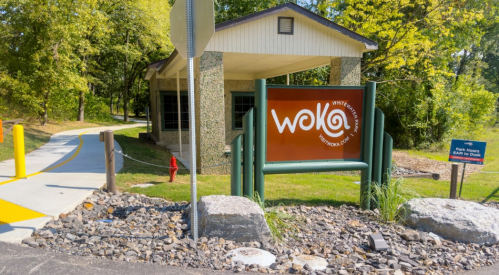 Sign for Woka, a visitor center, with a pathway and greenery in the background.