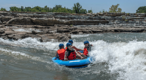 A group of five people in a blue raft navigating rapids on a river, surrounded by rocky terrain and greenery.