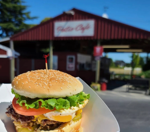 A close-up of a burger with lettuce, tomato, and a fried egg, in front of a red café building.