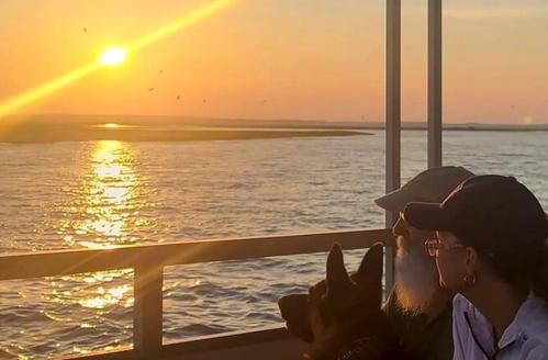 A person and a dog watch a sunset over the water from a boat, with warm colors reflecting on the surface.