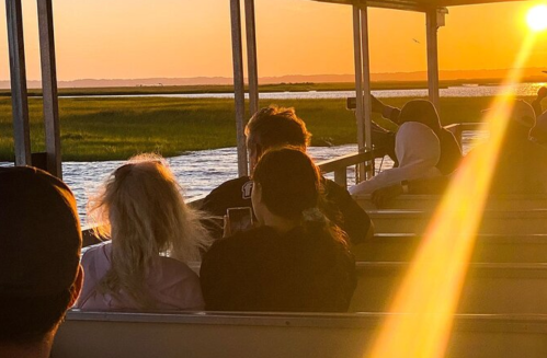 Silhouettes of people on a boat at sunset, with golden light reflecting on the water and lush greenery in the background.
