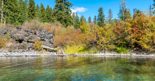 A serene river scene with clear water, rocky banks, and vibrant autumn foliage under a blue sky.