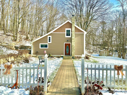 A modern house with a sloped roof, surrounded by snow and trees, with a stone pathway and white picket fence.