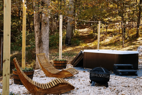 A cozy outdoor space with wooden lounge chairs, a hot tub, and a fire pit surrounded by trees and gravel.