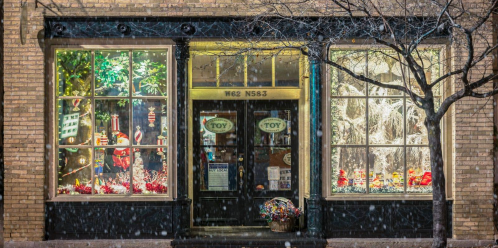 A festive toy store window decorated for winter, featuring colorful displays and a light snowfall outside.