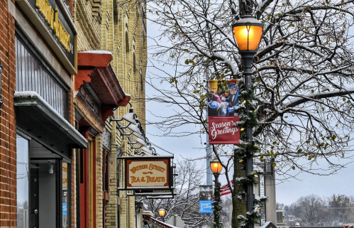 Snowy street scene featuring storefronts, a festive banner, and lampposts adorned with holiday decorations.