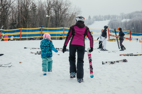 A woman and a child walk on snow, carrying skis, with other skiers and colorful barriers in the background.