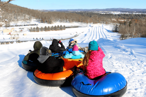 A group of people sitting on snow tubes, overlooking a snowy hill and tubing lanes in a winter landscape.