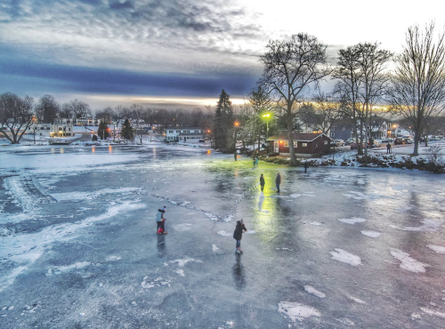 People ice skating on a frozen lake at sunset, surrounded by trees and houses, with a colorful sky above.