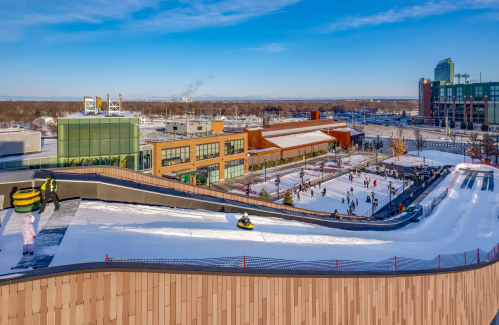 A snowy hill with people sledding, surrounded by buildings and a clear blue sky. Winter fun in an urban setting.